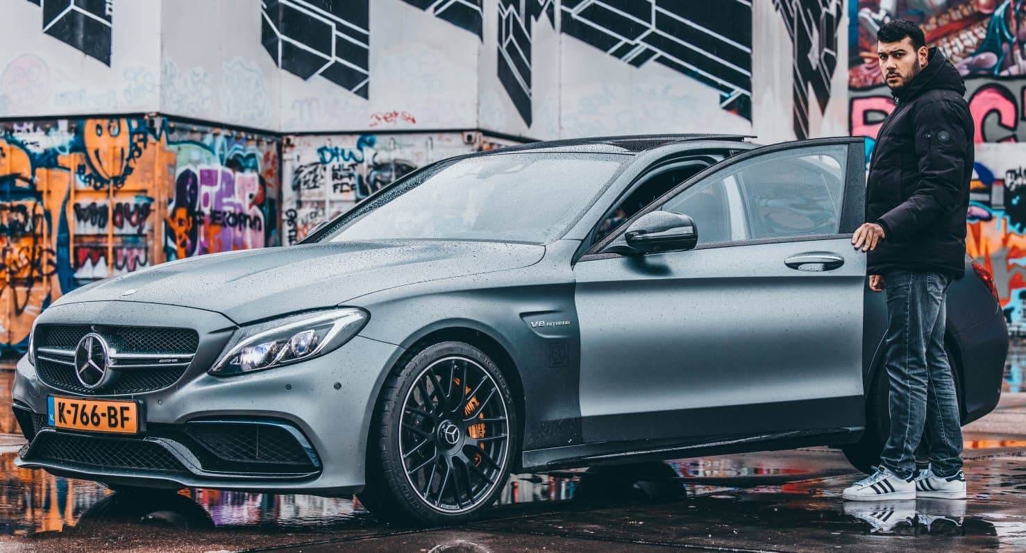 Emre naast zijn Mercedes C63 S AMG bij een graffitimuur